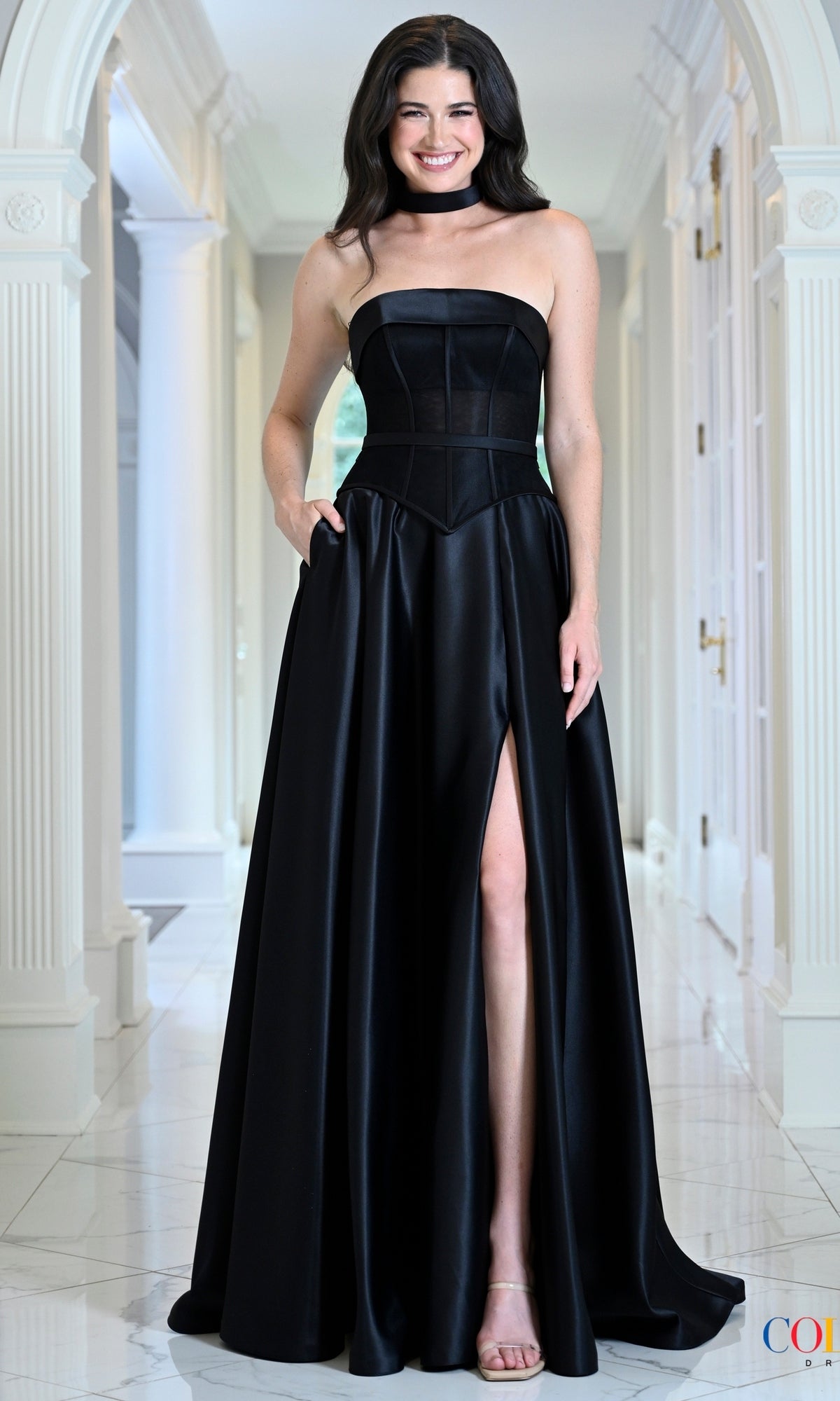 A smiling woman wears a Black Satin Prom Dress: Colors Dress 4056, featuring a strapless black corset bodice, a floor-length silhouette, and a side slit.